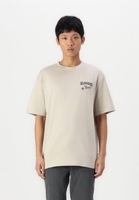 Camiseta beige de manga corta en algodón con cuello redondo, con un logo impreso en el lado izquierdo del pecho. Combinada con pantalones gris oscuro, de diseño sencillo.