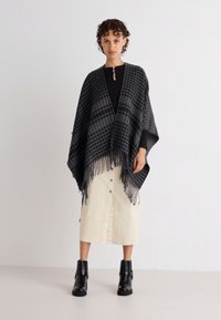 Schwarz-graues gemustertes Poncho mit Fransen, getragen über einem schwarzen Oberteil und einem cremfarbenen Rock, ergänzt durch schwarze Stiefeletten.