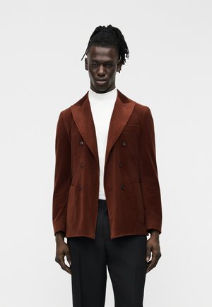 Mand med dreadlocks iført en brun dobbeltradet manchestermantel, hvid rullekrave og sorte bukser mod en enkel lys baggrund.