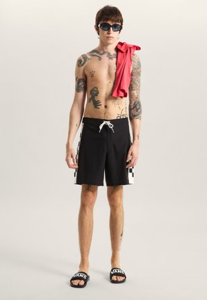 Pantaloncini da bagno neri con motivo a scacchi sui lati, abbinati a una maglietta rossa appoggiata sulla spalla e occhiali da sole scuri.