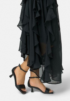 Pies con sandalias negras de tacón y tiras en el tobillo, con una falda negra de capas fluidas contra un fondo claro y liso.