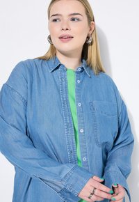 Camicia di jeans azzurro chiaro con maniche lunghe, chiusura a bottoni davanti e tasca sul petto, indossata sopra una maglietta verde brillante, che mostra un design casual.