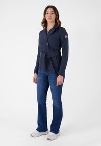 Giacca doppiopetto blu navy con cintura, realizzata in materiale liscio. Abbinata a jeans blu e scarpe da ginnastica bianche.