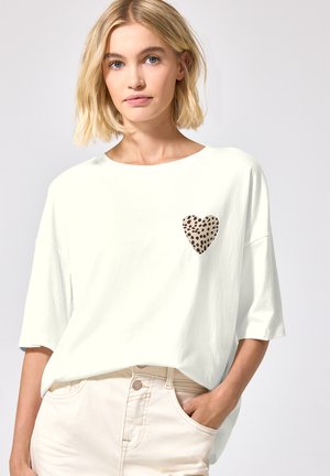 Femme blonde portant un T-shirt blanc ample à manches courtes avec un cœur imprimé léopard sur la poitrine gauche, main dans la poche d’un pantalon clair.