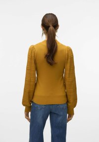 Senapsgul stickad tröja med texturerade, puffiga spetsärmar och figursytt liv. Matcher med löst sittande blå denimjeans, sedd från ryggen.