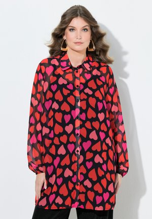 Kvinde iført sort bluse med lange ærmer med rødt og lyserødt hjerteprint, store guldringe øreringe, stående mod hvid baggrund.