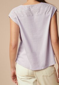 Haut lavande clair à manches courtes, présentant une broderie florale blanche complexe sur le plastron et une coupe décontractée, assorti à un pantalon crème.