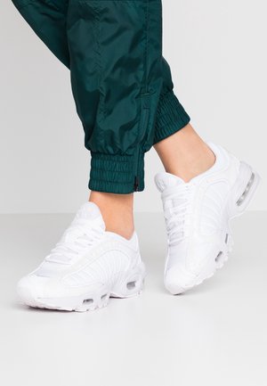 Osoba nosi białe sportowe trampki oraz ciemnozielone nylonowe spodnie dresowe z elastycznymi ściągaczami i zamkami przy kostkach.