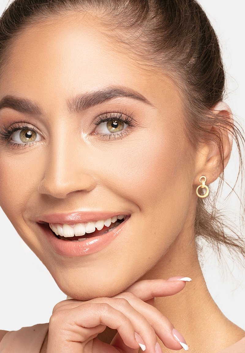 Orecchini a cerchio in oro con un design texturizzato, caratterizzati da una forma circolare liscia. La pelle è impeccabile con trucco naturale, sfondo chiaro.