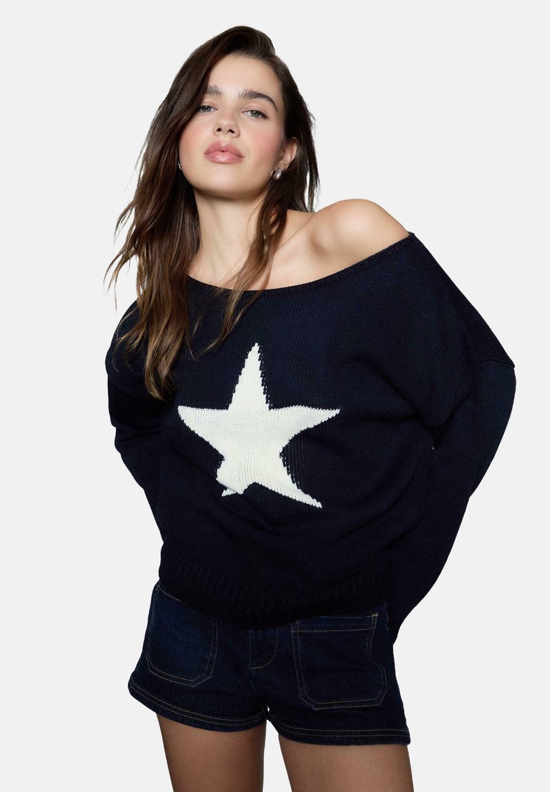 Marineblauer Strickpullover mit oversized Schnitt, der ein großes weißes Sternmuster auf der Vorderseite hat. Kombiniert mit dunklen Jeansshorts.