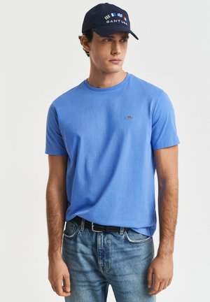 Uomo che indossa una camicia a maniche corte blu, jeans azzurri, cintura nera e cappello blu navy con disegni colorati di bandiere, che guarda di lato su uno sfondo semplice.