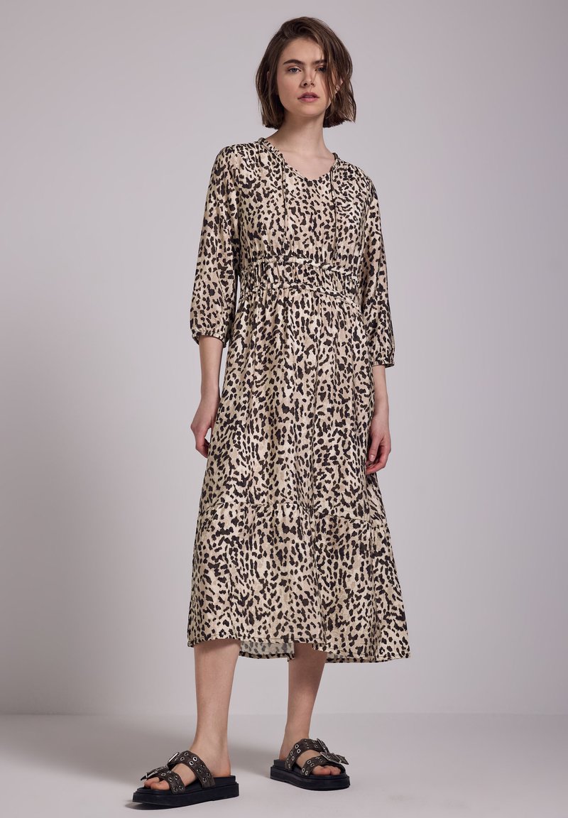 Donna in piedi con indosso un vestito leopardato beige e nero con maniche a tre quarti e sandali neri con fibbie.