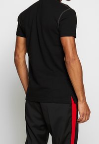 Zwart polo shirt met korte mouwen, voorzien van contrasterende stikdetails op de schouders, een rechte zoom en zijsplitten voor extra bewegingsvrijheid.