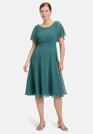 Robe en chiffon teal avec des manches courtes et fluides, taille resserrée et jupe évasée, assortie à des sandales à talons argentés.