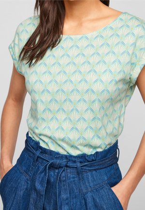 Kobieta ubrana w jasnoniebieską bluzkę z krótkim rękawem w wzory, wpuszczoną w wysokie, ciemnoniebieskie dżinsy z paskiem wiązanym w kokardę.