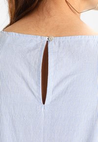 Haut à rayures bleu ciel et blanches, présentant un détail dos en keyhole maintenu par un bouton. Texture douce avec une coupe décontractée.