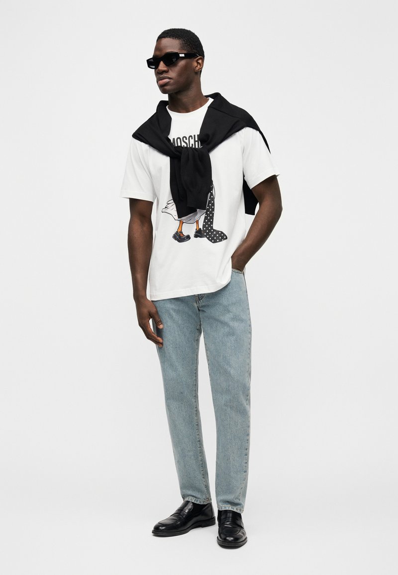 Weißes T-Shirt mit Grafikdruck, hellblaue Jeans, schwarze Slip-on Schuhe und ein schwarzer Pullover locker über die Schultern gelegt. Schwarze Sonnenbrille.