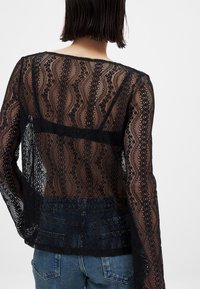 Zwarte kanten top met een golvend patroon, doorzichtige lange mouwen en een stevige taille, gecombineerd met blauwe denim jeans.