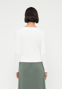 Άσπρο μακρυμάνικο τοπ με τετράγωνο λαιμόκοψη, συνδυασμένο με πράσινη φούστα. Το ύφασμα φαίνεται λείο και απαλό, με ελάχιστες ορατές ραφές.