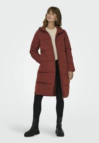 Manteau matelassé mi-long de couleur rouge foncé avec une capuche. Présente une fermeture éclair et des poches latérales, conçu en un matériau synthétique lisse.