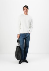 Hombre con camisa de manga larga blanca y jeans azules, sosteniendo un bolso de cuero negro, de pie frente a un fondo blanco liso.