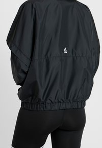 Chaqueta bomber negra de nailon con un corte relajado, cinturilla elástica y un pequeño logo en la parte de atrás. Textura suave, mangas cortas.