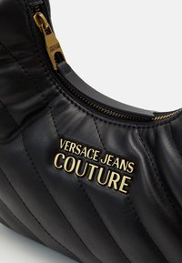 Černá kožená kabelka s prošívanou texturou, zlaté kovové doplňky s logem "VERSACE JEANS COUTURE" a zip detail podél horního okraje.