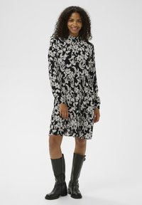 Must kleit valgete lille- ja paisleimustritega, pikkade varrukatega, kõrge kaelusega ja lõdva lõikega. Kandmiseks koos põlveni ulatuvate mustade saapadega.