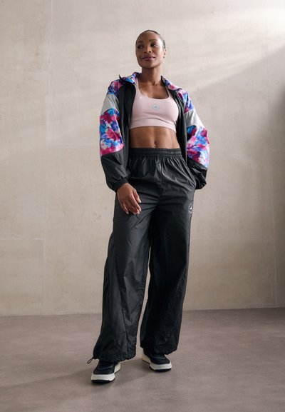 Tenue de sport comprenant une veste légère noire avec des manches en tie-dye coloré, un crop top rose clair et un pantalon noir ample.