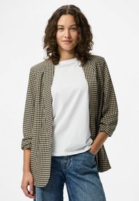 Mlada žena s kovrčavom kosom nosi bež i crni blazera u hođim uzorku preko bijele majice i plavih traperica, stojeći s jednom rukom u džepu.