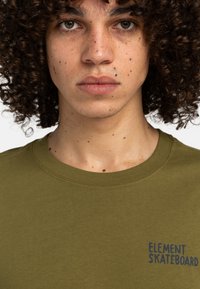 T-shirt en coton vert olive avec un col rond, présentant un logo "ELEMENT SKATEBOARD" bleu marine imprimé sur la poitrine gauche.