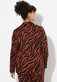 Blazer a strisce tigre in arancione e nero, con un design strutturato, maniche lunghe e una texture morbida. La vista posteriore mostra dettagli a coda.