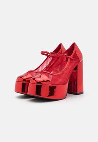 Tacones de plataforma rojos con un acabado de cuero sintético brillante, detalles de malla y tacones anchos de bloque. Presentan una punta redondeada y una correa en el frente.