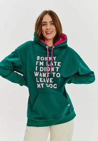 Zelená mikina s kapucí vyrobená z měkké látky, s růžovou podšívkou a velkým růžovým nápisem: "Omlouvám se, že spožuji, nechtěl/a jsem opustit svého psa." S nastavitelnými šňůrkami.