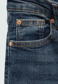 Temno modri kavbojke iz denima z oranžnim šivom, ki imajo sprednji žep s bakrenim zakovicom in gladko teksturo.