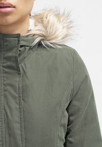 Oliwkowy parka z miękkim wykończeniem kaptura z sztucznego futra. Posiada zamek błyskawiczny i przeszycia na tkaninie. Miękka w dotyku tekstura.