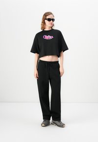 Kvinde iført sort cropped grafisk t-shirt, løse sorte bukser, grå sneakers og sorte solbriller, stående imod en hvid baggrund.