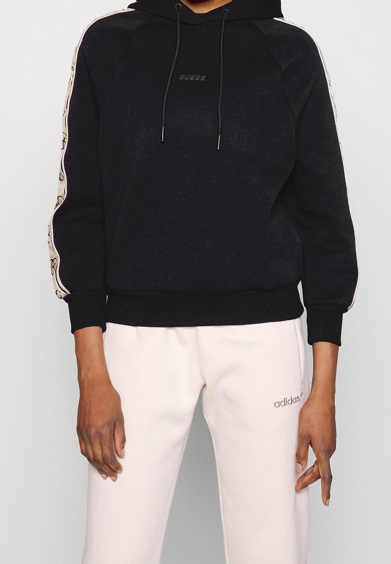 Personne portant un sweat à capuche noir Guess avec des rayures à motifs sur les manches et un pantalon de survêtement blanc Adidas, debout devant un fond clair.