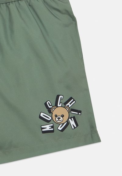 Πράσινα σορτς με ελαστική μέση διαθέτουν γραφικό αρκούδας και έντονο κείμενο σε μαύρο και άσπρο. Το ύφασμα είναι λείο με ελάχιστη γυαλάδα.