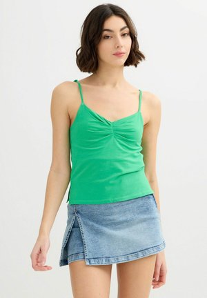 Mujer con cabello corto y oscuro, vestida con una camiseta verde drapeada y una minifalda de mezclilla azul, sobre un fondo blanco.