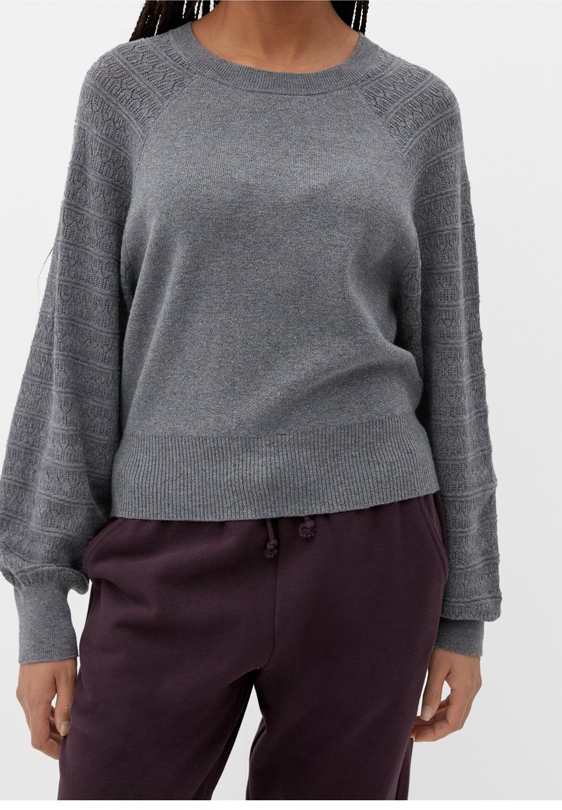 Femme portant un pull en maille gris avec des manches ballon texturées et un ourlet côtelé, associé à un pantalon violet foncé à cordon.
