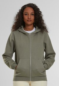 Sudadera verde con cremallera hecha de tela suave, con capucha, bolsillos frontales y puños de canalé con una textura lisa.