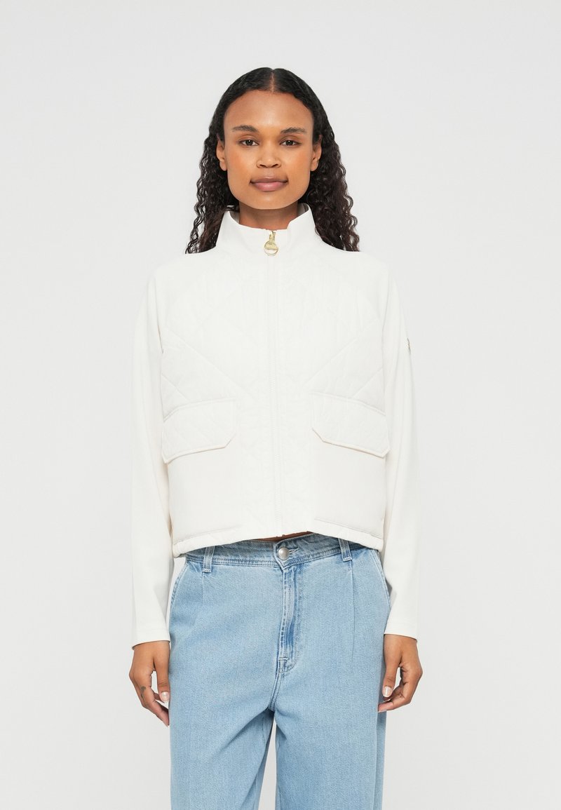 Jaqueta branca quiltada cropped com gola alta, fecho éclair frontal e dois grandes bolsos no peito. Possui uma textura suave e detalhes em metal dourado.