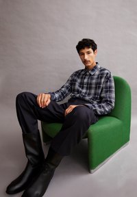 Geruit marineblauw en witte button-up shirt, donkere broek en kniehoge zwarte laarzen, zittend op een levendig groene moderne stoel tegen een grijze achtergrond.