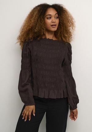 Blouse marron smockée avec de longues manches bouffantes et un ourlet volants. Le tissu a une texture lisse avec des détails froncés partout.