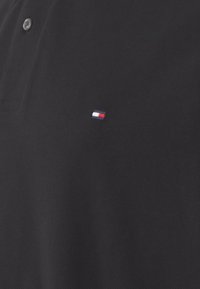 Polo noir en tissu texturé avec patte de boutonnage à trois boutons. Petit logo brodé en rouge, blanc et bleu sur la poitrine.