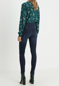Jeans ajustados de color azul oscuro con una cintura ceñida, combinados con una blusa verde con un patrón floral blanco, y luciendo botas de tacón bloque verde.