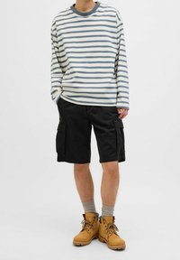 Pasiasta koszula z długim rękawem w kolorze kremowym i szarym, połączona z czarnymi szortami cargo. Na nogach brązowe buty trekkingowe i szare skarpetki.