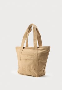 Sac fourre-tout en toile beige avec deux poignées et une poche avant, debout verticalement sur un fond blanc.