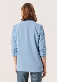 Blazer azul claro con hombros estructurados, puños enrollados, tejido suave y un diseño liso, visto desde la parte posterior.